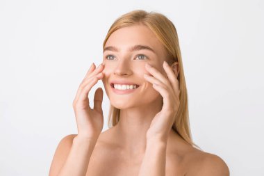 Cilt bakımı. Neşeli Sarışın Kadın Yüze Dokunan Yumuşak Tenli Beyaz Stüdyo Arkaplanına Bakarken Krem Uygulama. Güzellik ve Kozmetik