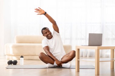 Çevrimiçi Antrenman Konsepti. Gülümseyen, gülümseyen siyah adam oturma odasında antrenman yapıyor, vücudu esniyor. Yoga Mat 'te oturan mutlu Afrikalı Amerikalı adam dizüstü bilgisayar kullanıyor, özel ders izliyor ve ekranlara bakıyor.