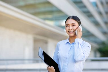 İş İletişim Konsepti. Mutlu Koreli genç iş kadını cep telefonuyla konuşuyor ve gülümsüyor, dışarıda dizüstü bilgisayar ve tablet bilgisayarla bekliyor.