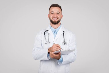 Beyaz önlüklü ve steteskoplu, kendinden emin genç bir erkek doktor. Elinde bir pano var, notlar yazıyor. Tarafsız gri arka planda hoş bir gülümsemeyle.