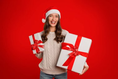 Neşeli bayan kırmızı stüdyo arka planına karşı iki güzel paketlenmiş Xmas kutusunu tutuyor. Kış tatillerinin merak ve cazibesini ifade ediyor. Neşeyi kutluyor, kameraya gülümsüyor.