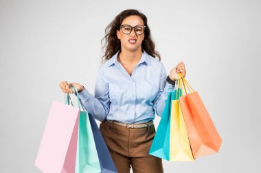 Resmi giyinmiş yorgun genç Avrupalı kadın gri arka plandaki stüdyo arka planında izole edilmiş bir sürü alışveriş çantası taşıyor. Büyük indirim, alışveriş merkezinden kıyafet alma sorunları, moda.