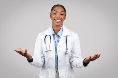 Gülümseyen Afrikalı Amerikalı kadın doktor ellerinde boş bir alan tutuyor, gri arka planda, stüdyoda. Tıbbi yardım, modern sağlık hizmeti tavsiyesi, seçim, reklam ve teklif