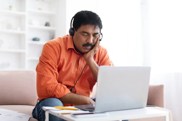 Tired middle aged indian man with moustache wearing casual outfit working from home, sitting on couch in living room with closed eyes, using headset and laptop, feeling sleepy, copy space