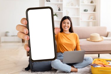 Sıcak seyahat teklifi. Mutlu genç Asyalı kadın gezgin evde oturmuş dizüstü bilgisayar kullanıyor ve beyaz ekranlı akıllı telefonu gösteriyor. Reklam için maket yapıyor, bavul hazırlıyor.