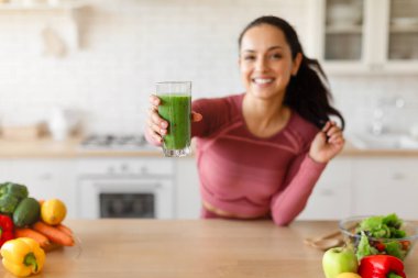 Spor salonundaki kadın mutfağında duruyor ve kameraya taze yapılmış yeşil smoothie ikram ediyor. Gülümsüyor ve sağlık için kilo kaybı yemeği öneriyor. Cam üzerine seçici odaklanma