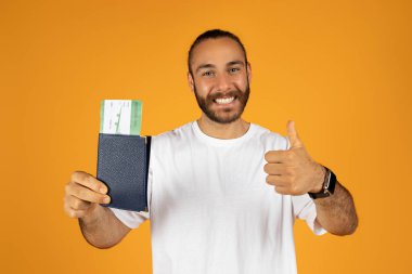 Beyaz tişörtlü memnun Avrupalı adam pasaport ve bilet gösteriyor, yaşam tarzını seviyor, turuncu stüdyo arka planında izole edilmiş. Turizm, yaz tatili, seyahat ve macera