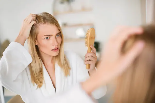 Worried Young Blonde Woman Checking Her Hair Roots Standing Near Mirror At Home, Having Dandruff Problem. Upset Lady In Bathrobe Suffering From Hair Loss And Flaky Scalp Issue. Selective Focus