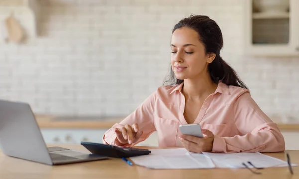 stock image Focused young woman in a pink blouse, working at a wooden desk with a laptop, browsing her smartphone, with papers and pen nearby in a bright, contemporary kitchen setting, free space