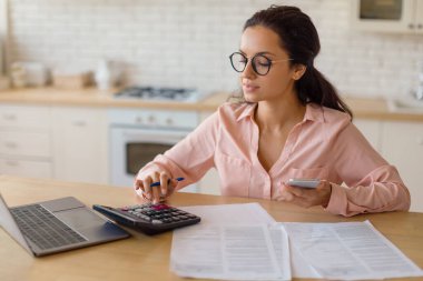 Ahşap mutfak masasında dokümanlarla çevrili akıllı telefon ve hesap makinesi kullanan gözlüklü genç bir kadın. Modern bir konut ortamında finansal görevler üzerinde çalışıyor gibi görünüyor..