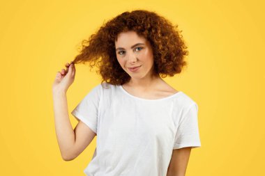 Beyaz tişörtlü, kıvırcık saçlı pozitif genç bir kadın saçlarına dokunuyor, boş zamanların tadını çıkarıyor, sarı arka planda, stüdyoda izole edilmiş. Öğrenciler flört etmeyi, aşkı, yaşam tarzını ve romantik duyguları severler.
