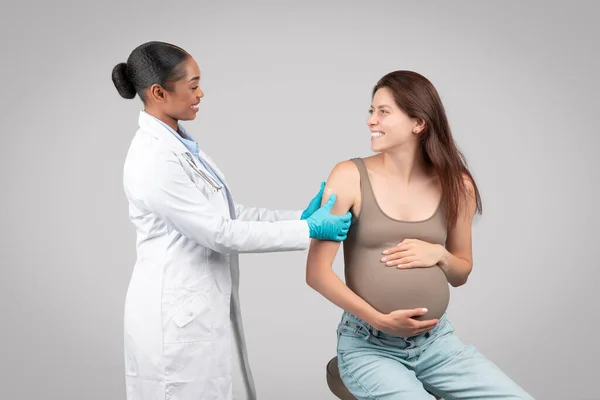 Cheerful black woman doctor make injection to pregnant patient during pregnancy, puts plaster on hand, isolated on gray background studio. Vaccination in clinic, health care, expecting baby
