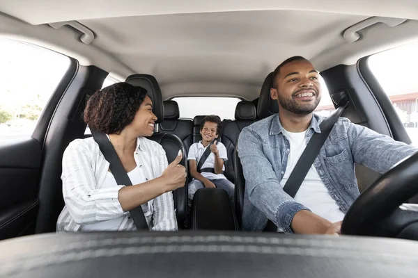 一路顺风快乐的黑人家庭 年轻的父母和儿子一起享受汽车旅行 非裔美国母亲用安全带看着她的孩子 露出大拇指 父亲驾驶汽车 面带微笑 — 图库照片