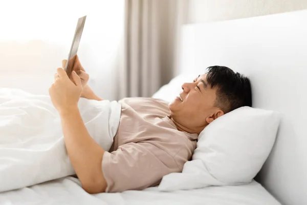 Happy Middle Aged Chinese Man Using Modern Digital Tablet While Relaxing In Bed, Resting In Bedroom With Tab Computer, Making Online Shopping Or Browsing Internet At Home, Side View