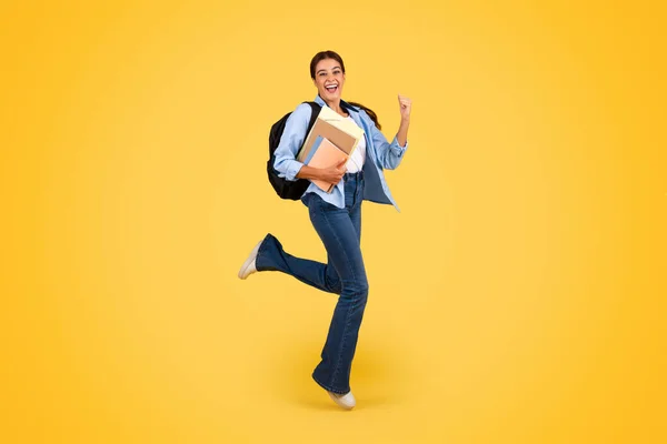 快乐的年轻的欧洲姑娘 背着背包和书本跳着 空气冰冷 享受着学生的生活方式 被隔离在黄色的工作室背景下 教育和学习 — 图库照片