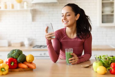 Diyet uygulaması. Spor kıyafetleri giymiş bir kadın cep telefonu kullanıyor ve mutfakta kilo verme içeceği içiyor. Sağlıklı beslenme sitesi ve zayıflatma uygulaması reklamı