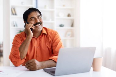 Orta yaşlı Hintli bir adam cep telefonuyla konuşuyor, dizüstü bilgisayarla masada oturuyor ve ev ofisinden internete girip boş alana bakıyor. İş ve mobil iletişim kavramı