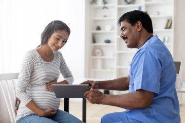 Klinikte hamile bir kadına dijital tablet gösteren olgun Hintli doktor. Modern hastanede hamile bir kadına doğum öncesi bilgi veren üniformalı profesyonel jinekolog.