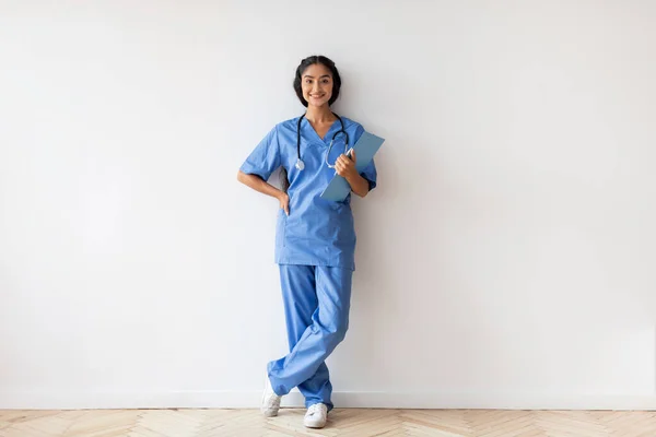 Full Length Of Smiling Young Indian Doctor Woman In Uniform With Clipboard In Hands Posing Against White Wall In Clinic, Happy Female Physician With Stethoscope Looking At Camera, Copy Space