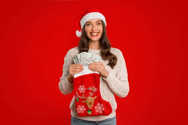 寛大なクリスマスプレゼント サンタハットで若い女性 キャッシュマネーを保持し Xmasストッキング ソックス スタンド レッドスタジオ背景 笑顔に カメラ 金融プレゼントを受け取る — ストック写真