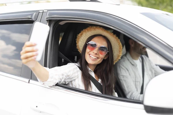 Stylish happy young woman taking selfie on phone during car ride with her husband or boyfriend, wearing sunglasses and wicker hat. Travel blogger sharing nice photo while have vacation