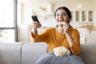 Neşeli genç Hintli kadın TV izliyor ve evde patlamış mısır yiyor, oturma odasındaki koltukta dinlenirken uzaktan kumandayla kanal değiştiriyor, hafta sonları boş zamanların tadını çıkarıyor,