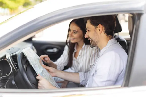 Road Trip Concept. Side view from open window of smiling man and lady holding and looking at paper map sitting inside car, choosing location, have conversation, travelling together by auto