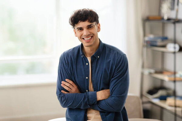 Портрет красивого арабского бизнесмена, стоящего со сложенными руками возле стола в офисе, уверенного молодого человека с Ближнего Востока, одетого в повседневную одежду, позирующего в помещении, улыбающегося перед камерой