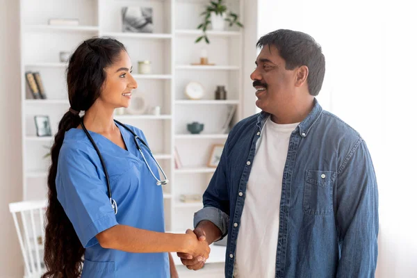 Thankful mature indian man shaking hands with female doctor in clinic, happy young physician lady in blue uniform greeting male patient in hospital, handshaking after successful treatment,