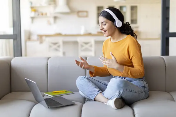 Video Call. Smiling Young Indian Woman In Headphones Teleconferencing On Laptop At Home, Happy Eastern Female Talking And Gesturing At Computer Web Camera While Sitting On Couch, Free Space