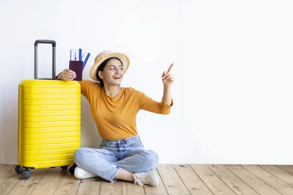Excited Young Indian Woman With Suitcase And Flight Tickets Pointing Aside At Copy Space With Finger, Happy Female In Straw Hat Ready For Vacation Travel, Sitting On Floor Near White Wall Indoors