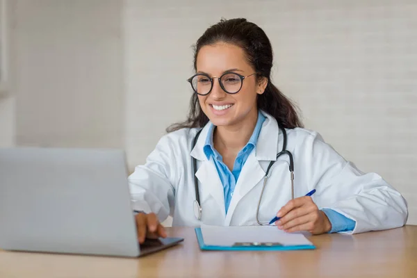 Cheerful female doctor wearing glasses, sitting at desk with laptop, holding clipboard and pen, showcasing a pleasant demeanor while in a professional medical setting.
