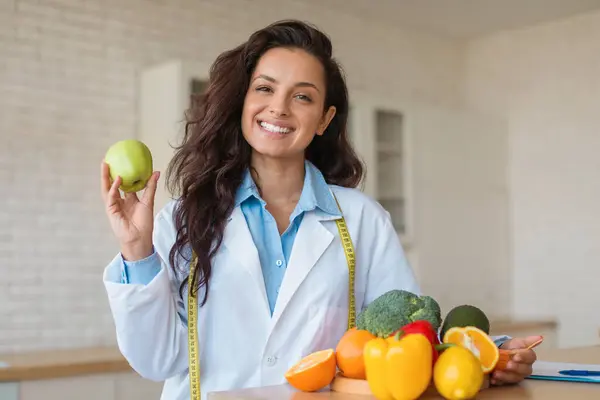 Joyful nutritionist holds green apple, standing next to table of fresh fruits and veggies, embodying the essence of a healthy diet and wellness