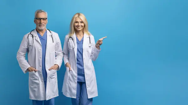 Elderly european doc man and woman posing together, experienced doctors wearing lab coats, standing over blue studio background, pointing at copy space, panorama