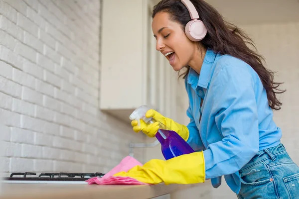 身穿蓝色衬衫 头戴黄色手套的充满热情的年轻女子一边拿着喷雾器和布来唱歌 一边清扫厨房柜台 一边用粉色耳机欣赏着她的音乐 — 图库照片