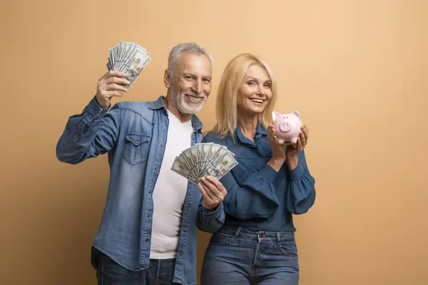 Wealthy rich happy retired man and woman beautiful senior couple posing with money dollar banknotes and piggy bank isolated on beige background, showing their savings, deposit cash, copy space