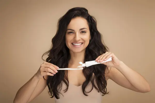 Cheerful brunette long-haired young woman holding wooden or bamboo organic eco-friendly teeth brush and toothpaste, showing her perfect teeth, isolated on beige background. Dental care concept