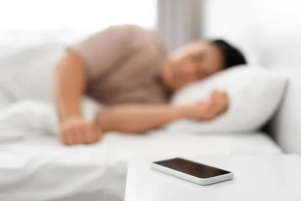 Early awakening concept. Asian man sleeping through ringing alarm on phone in the morning, lying in bed, selective focus on smartphone. Oversleeping, time to wake-up and get up. Shallow depth