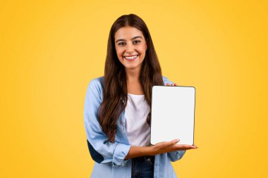 Pozitif Avrupalı genç bayan öğrenci ekranı boş, sarı arka planda, stüdyoda izole bir tablet gösteriyor. Uzaktan tavsiye, eğitim ve çevrimiçi ders, dikkat kursları uygulaması