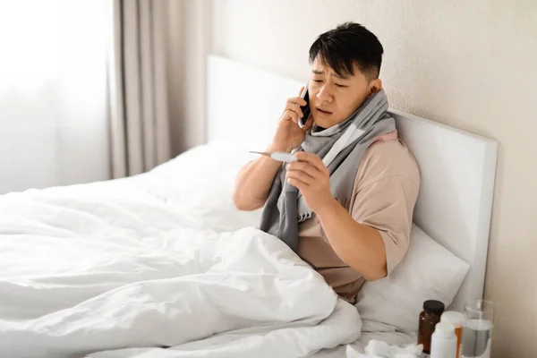 Online Consultation. Sick Mature Asian Man Wrapped In Scarf Holding Thermometer And Calling Doctor On Phone While Sitting In Bed At Home, Ill Chinese Man Suffering Cold Or Fever, Free Space
