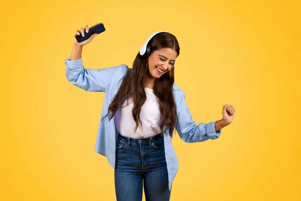 Cheerful young student woman with smartphone, headphones, dance, enjoy music, lifestyle, isolated on yellow background. Rest, relax at spare time, app for listen audio app