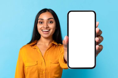 Hindu milenyum kadını elinde boş ekranlı büyük bir akıllı telefon tutuyor, mavi stüdyo arka planında uygulama modeli gösteriyor. Telefon kullanıcısı reklam dijital iletişim, modern aygıt şablonu