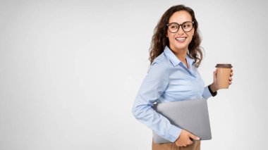 Gülümseyen Avrupalı genç bayan profesyonel öğretmen dizüstü bilgisayar ve kahve servisi yapıyor, boş alana bakıyor, gri stüdyo arka planında izole edilmiş. İş, uzaktan iş, mola, reklam ve teklif