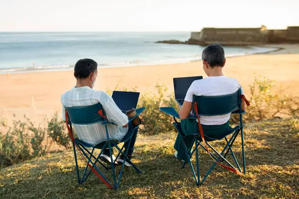 Seaside Office. Mature couple immersed in online tasks typing and browsing on their laptops, sitting comfortably on the serene oceanside shore, embracing the modern concept of remote work