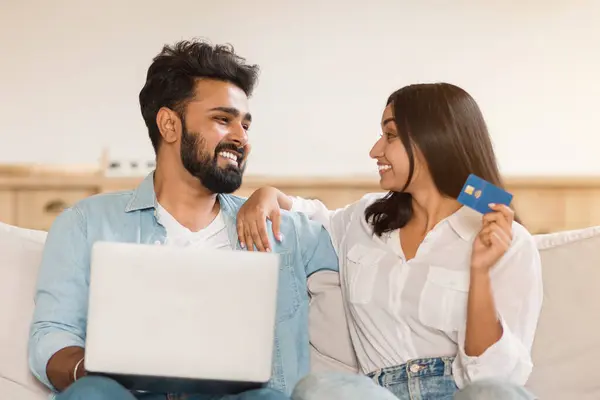 Young indian couple enjoys time together, man using laptop, woman holding credit card, looking at each other and smiling, resting on sofa at home