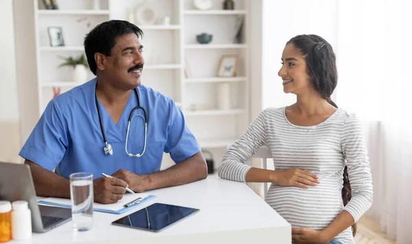 Young Pregnant Eastern Woman Getting Medical Consultation With Gynecologist In Clinic, Indian Mature Doctor Male Talking To Expectant Female Patient During Meeting At Office, Lady Embracing Belly