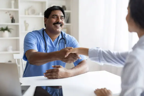 Friendly mature indian doctor man smiling and shaking female patient hand, smiling eastern therapist male greeting young woman at his clinic, outstretching hand, welcoming in office, copy space