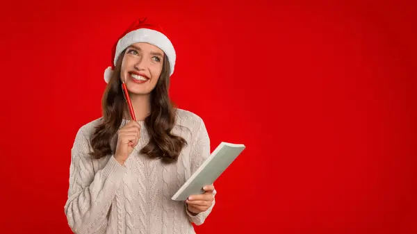 Christmas, New Year Wishes And Plans. Happy Lady In Santa Hat Posing With Pen And Notebook, Planning Upcoming Year, Writing Wishlist Letter To Santa Claus Over Red Background. Panorama, Copy Space