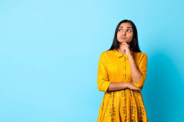 Bir düşüneyim. Altın işlemeli geleneksel sarı elbiseli düşünceli genç Hintli bayan, mavi stüdyo arkaplanı karşısında düşüncelere dalmış poz veriyor. Reklam için boşluk kopyala