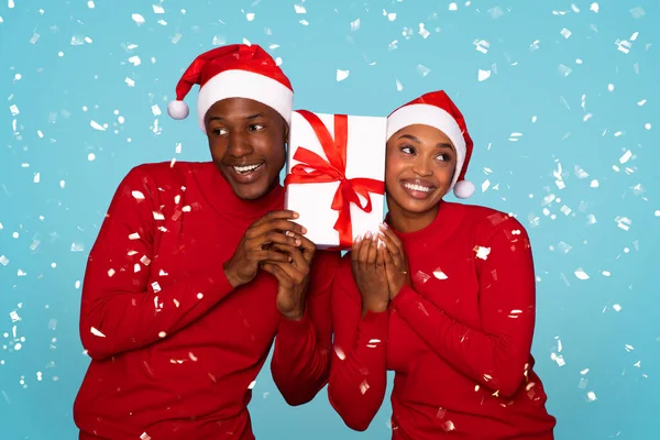Best Xmas Gifts. Black Woman And Man Wearing Santa Hats, Posing With Christmas Gift While Celebrating Family Holiday Against Blue Backdrop, Standing Under Falling Confetti